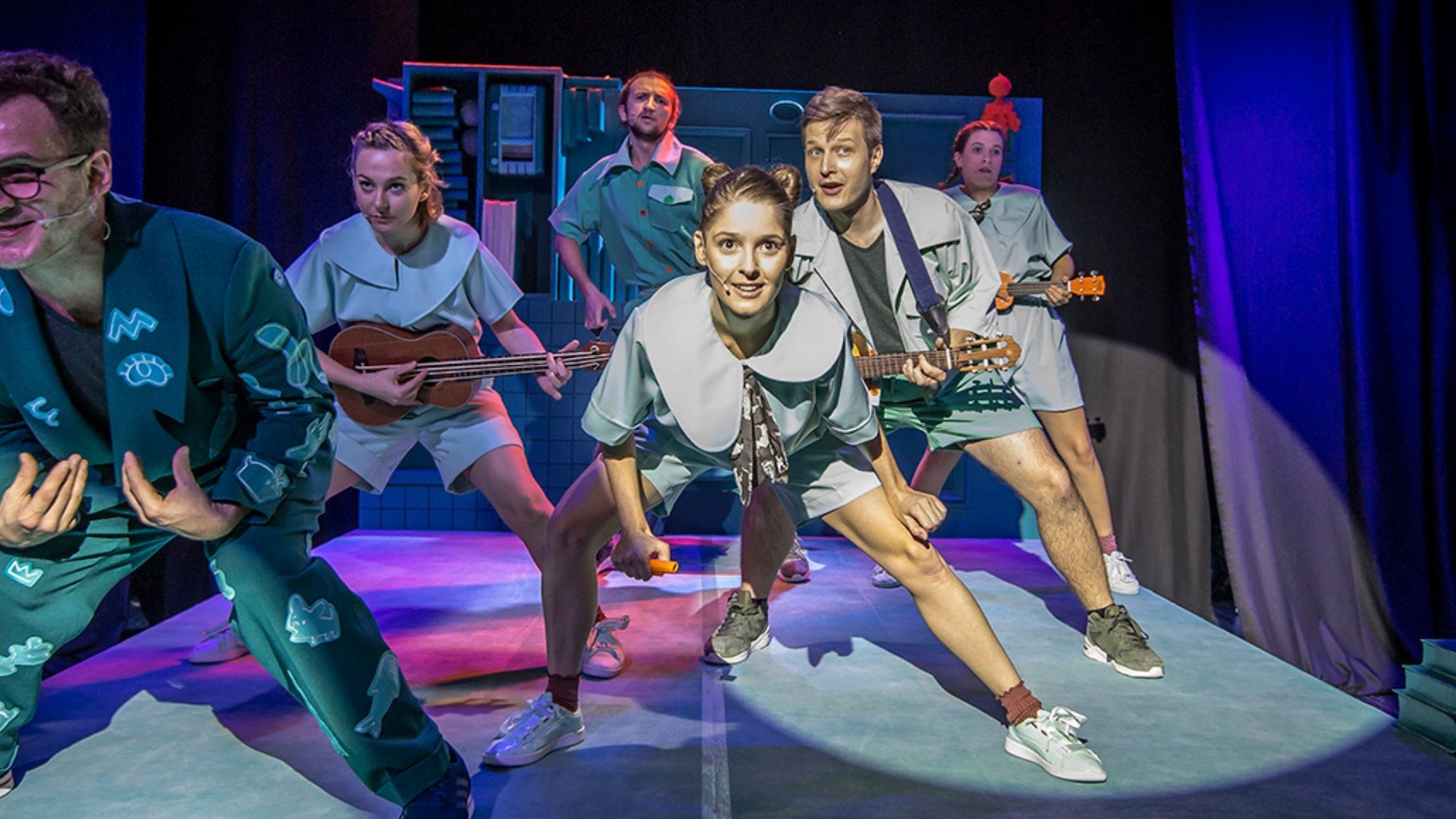 Scena zbiorowa. Mała grupa aktorów, część z nich trzyma w ręku małe gitary. Stoją w lekkim rozkroku wykonując choreografię.