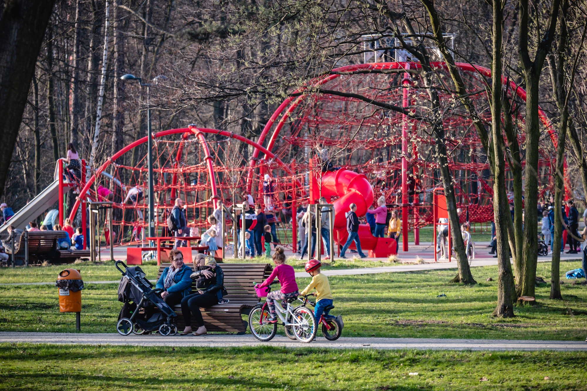 mali rowerzyści jadą po ścieżce, za nimi na lawce siedzą mamy, w tle na linarium bawią się licznie dzieci