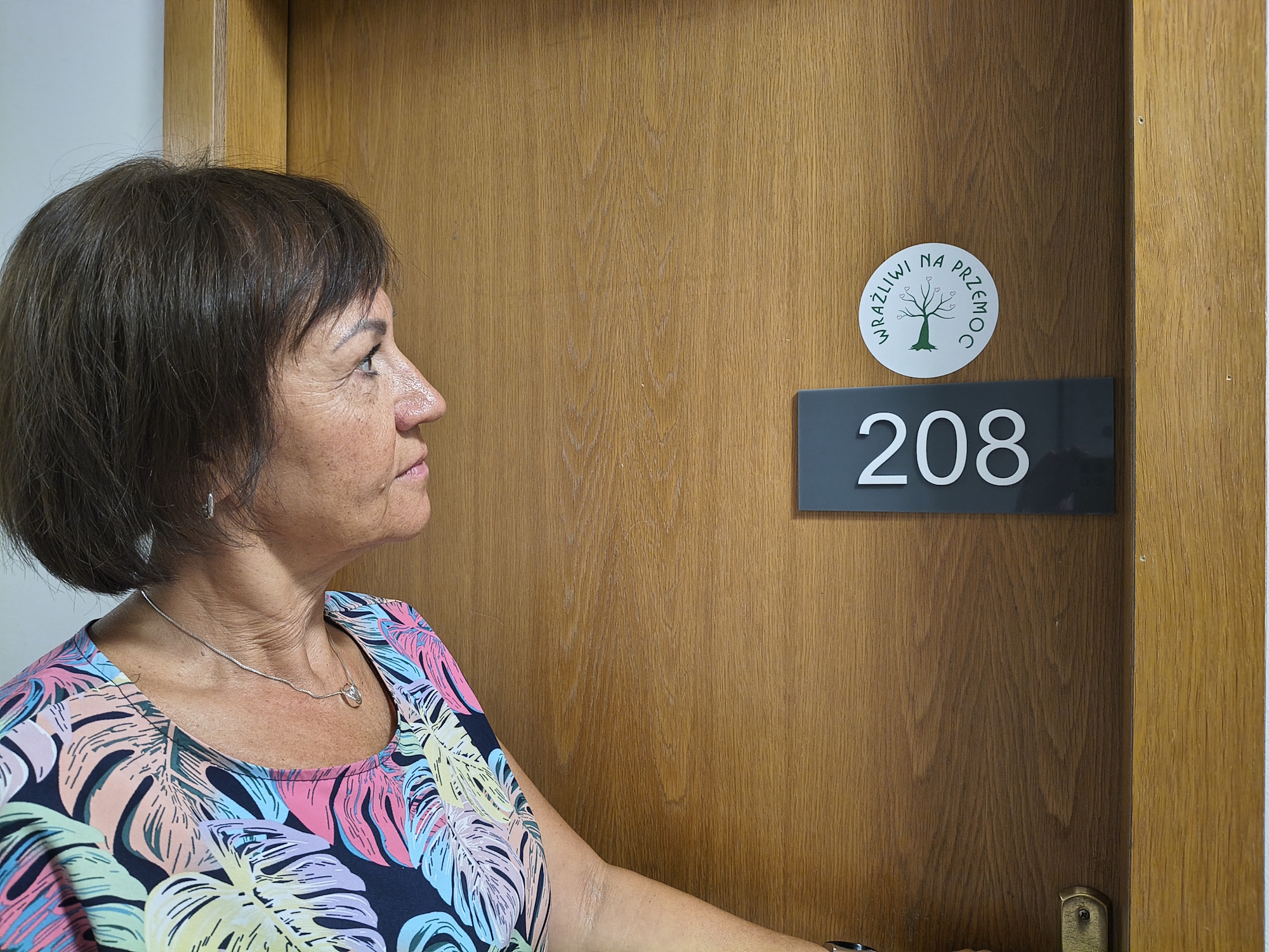 kobieta otwiera drzwi pokoju 208 , na drzwiach naklejka Wrażliwi na przemoc