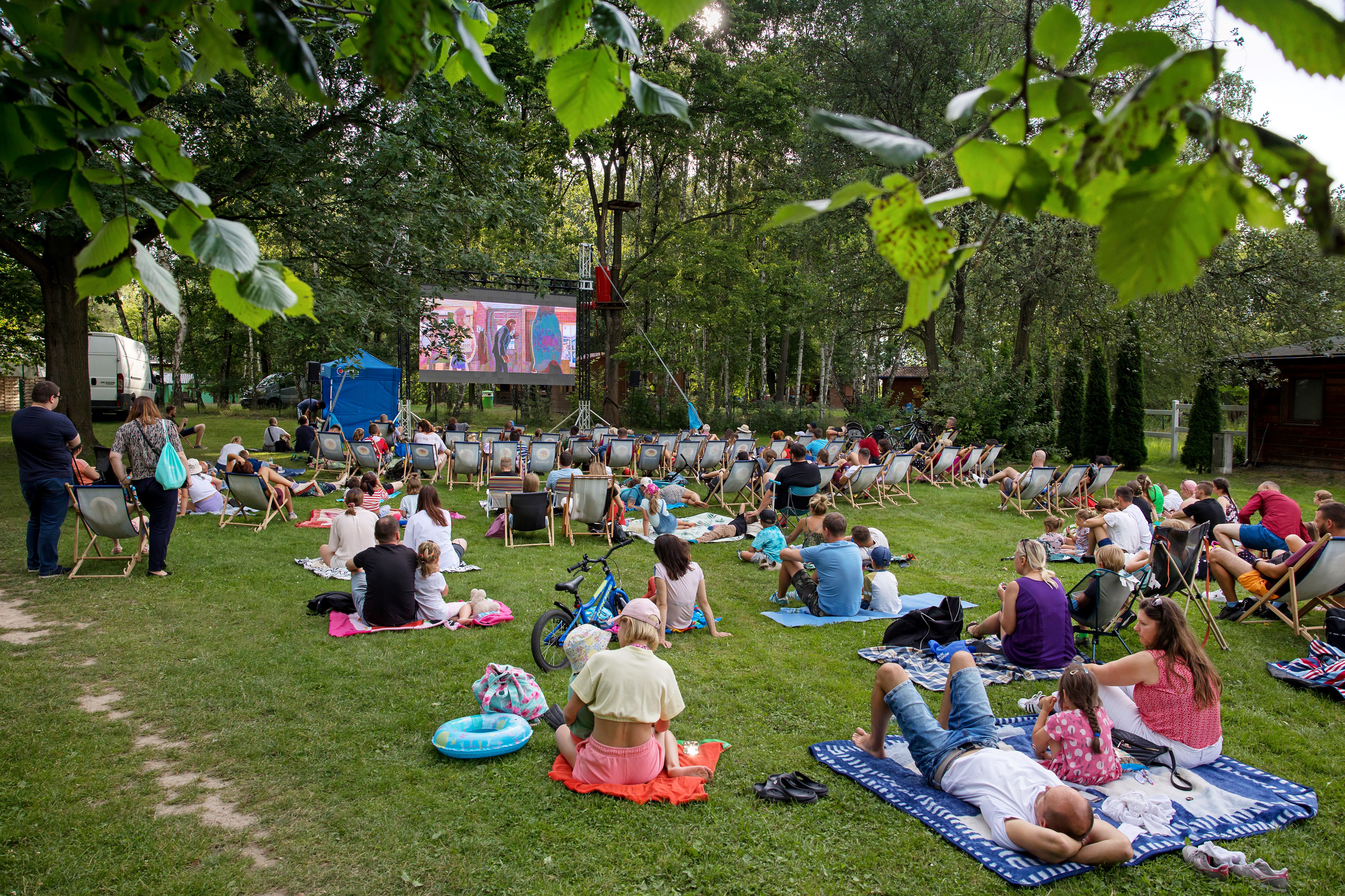 Ludzie siedzą na lezakach i kocach, oglądają film na ekranie w parku