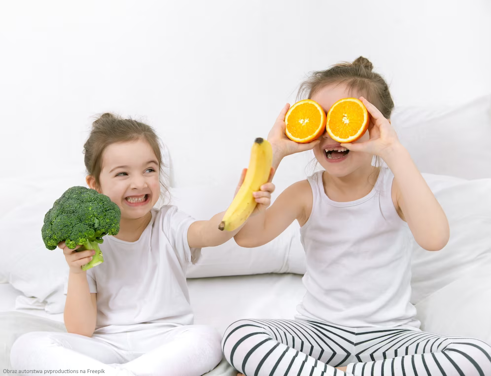 Dwie wesołe dziewczynki trzymające warzywa i owoce - brokuły, banany, pomarańcze.