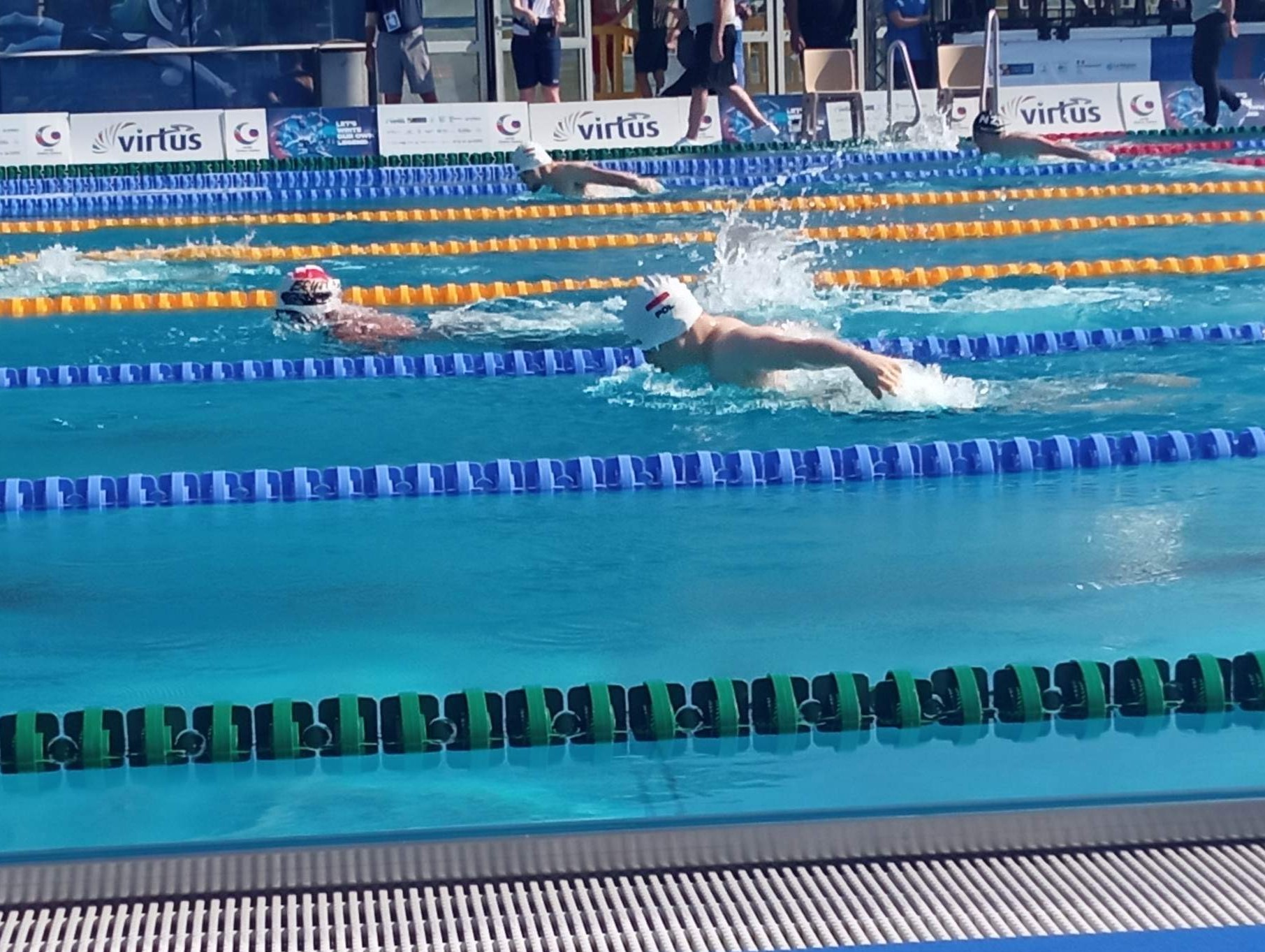 Pływacy rywalizują w wyścigu na basenie. W wodzie widocznych jest czterech zawodników, w tym dwóch ma na czepkach flagę Polski.