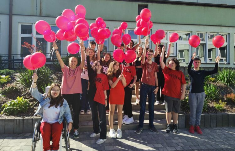 Grupa uśmiechniętej młodzieży, w tym dziewczyna poruszająca się na wózku, stoi przed szkołą. Większość ma na sobie czerwone elementy odzieży - koszulki, sukienki, spodnie. Nad głowami trzymają czerwone balony na patykach.