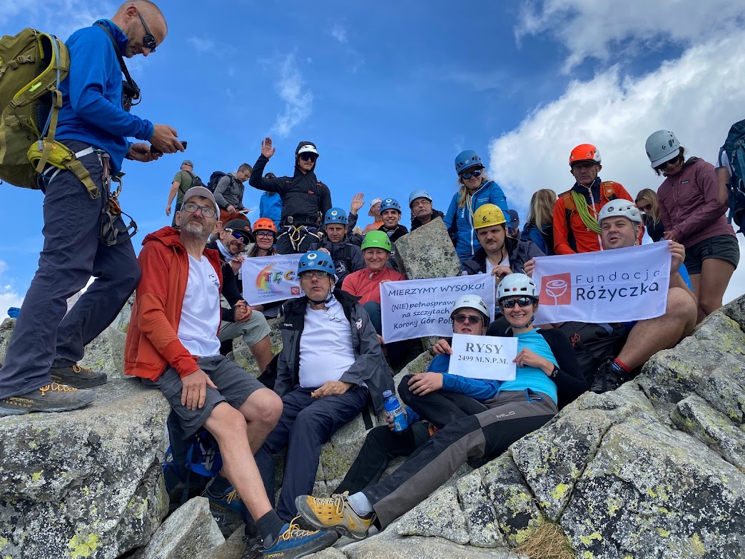 Grupa ludzi siedząca na skałach w górach,część osób w kaskach, w rękach trzymający napisy RYSY 2499 m n.pm., WTZ TĘCZA, Fundacja Różyczka, Mierzymy Wysoko, w tle niebiesko niebo.