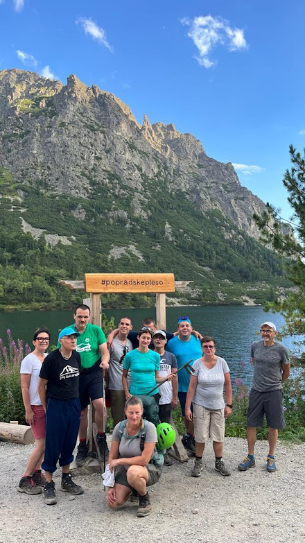 Grupa ludzi na tle bramki drewnianej z napisem Popradskepleso, z tyłu jezioro górskie, za jeziorem strome szczyty górskie, w tle niebieskie niebo.