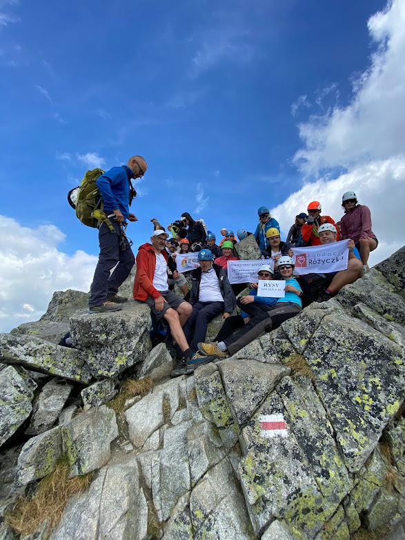 Grupa ludzi siedząca na skałach w górach, część osób w kaskach, w rękach trzymający napisy RYSY 2499 m n.pm., WTZ TĘCZA, Fundacja Różyczka, Mierzymy Wysoko, w tle niebiesko niebo, na pierwszym planie na skale znak czerwonego szlaku górskiego.