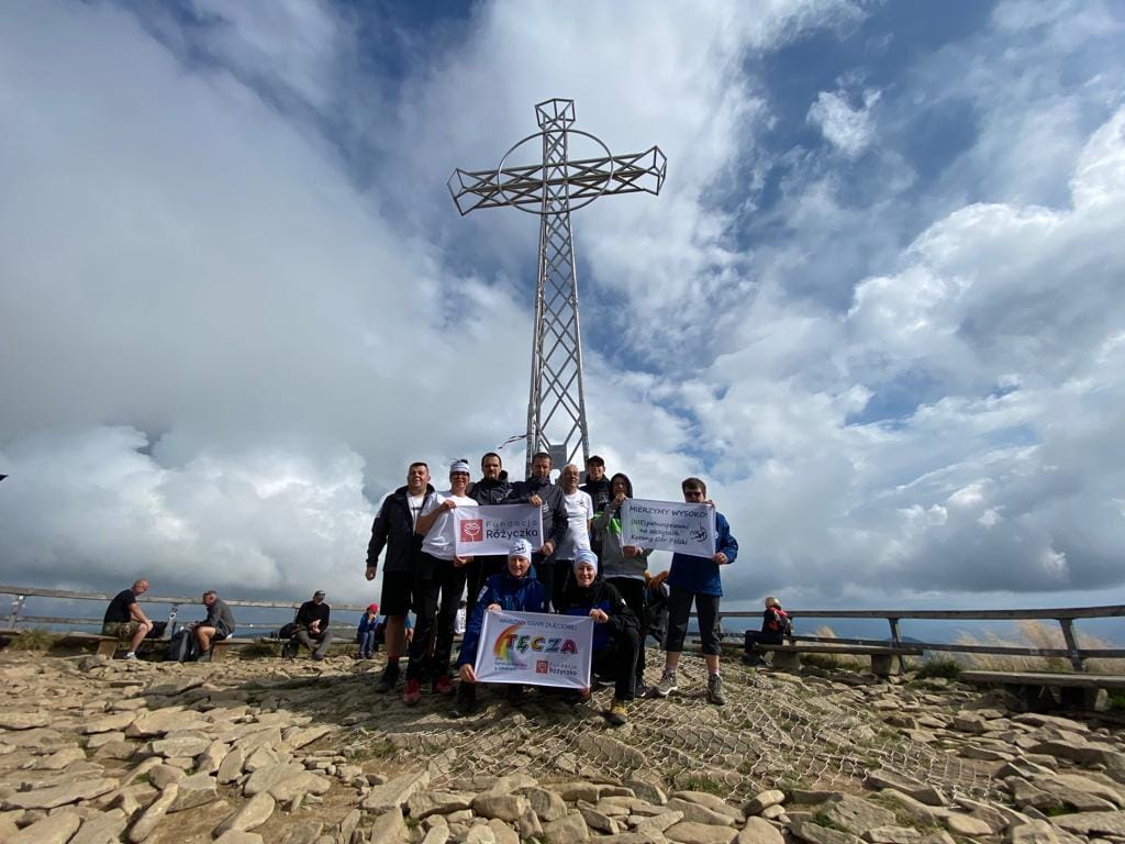 Grupa ludzi stoi przed metalowym dużym krzyżem na szczycie góry Tarnica, w rękach trzymają napisy, WTZ TĘCZA, Fundacja Różyczka, Mierzymy Wysoko, w tle niebo z chmurami.