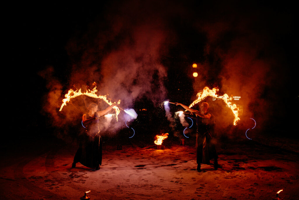 Dwie tancerki w czarnych strojach, w dłoniach trzymają płonące flary, za nimi płonące konstrukcje. Tańczą na śniegu, jest ciemno, jedynie ogień rozświetla noc.