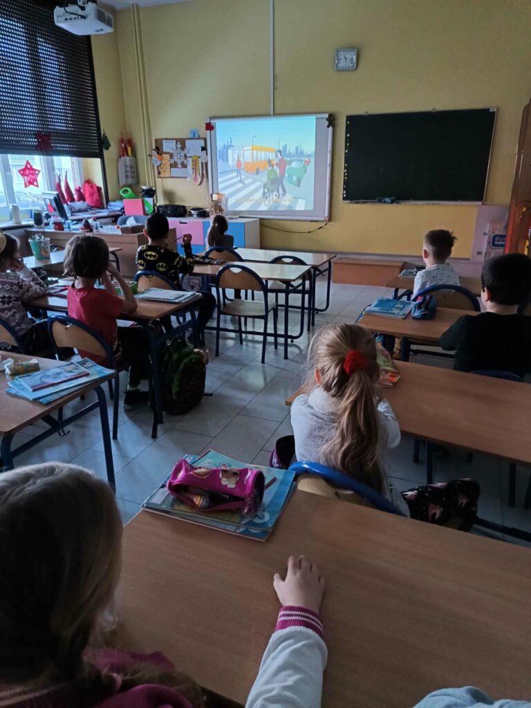 Widoczne od tyłu dzieci w sali szkolnej oglądające na dużym ekranie animację, na której osoba na wózku przejeżdża przez przejście dla pieszych.