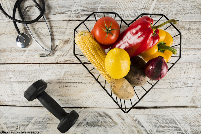 Stetoskop, czarne hantle i metalowy koszyk w kształcie serca wypełniony owocami i warzywami