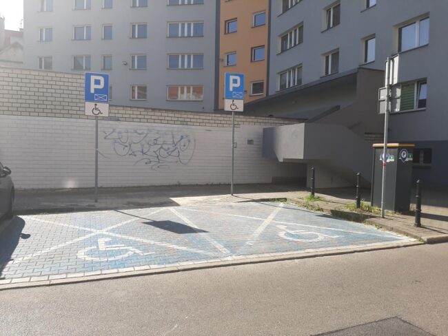 Dwa miejsca parkingowe oznakowane jako miejsca dla osób z niepełnosprawnością. W tle bloki mieszkalne.